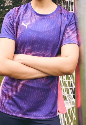 Osoba ubrana w fioletowo-różową sportową koszulkę Puma stoi ze skrzyżowanymi ramionami przed bramką na trawiastym boisku.