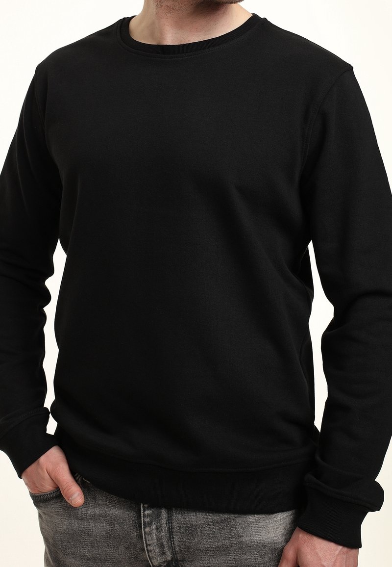 Homme portant un pull à col rond noir uni à manches longues avec poignets et ourlet côtelés, jean gris, main rentrée dans la poche.