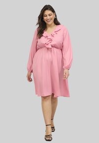 Rosa Chiffonkleid mit langen Puffärmeln, einem Rüschenausschnitt in V-Form und einem elastischen Bund, der knapp oberhalb des Knies endet. Kombiniert mit schwarzen Riemchensandalen.