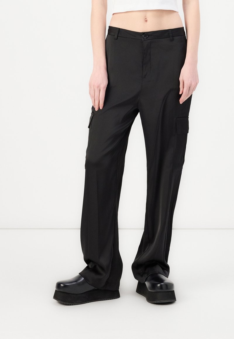 Persona con pantalones cargo negros holgados con bolsillos laterales y zapatos plataforma negros, top blanco corto, de pie sobre un suelo y fondo blancos.