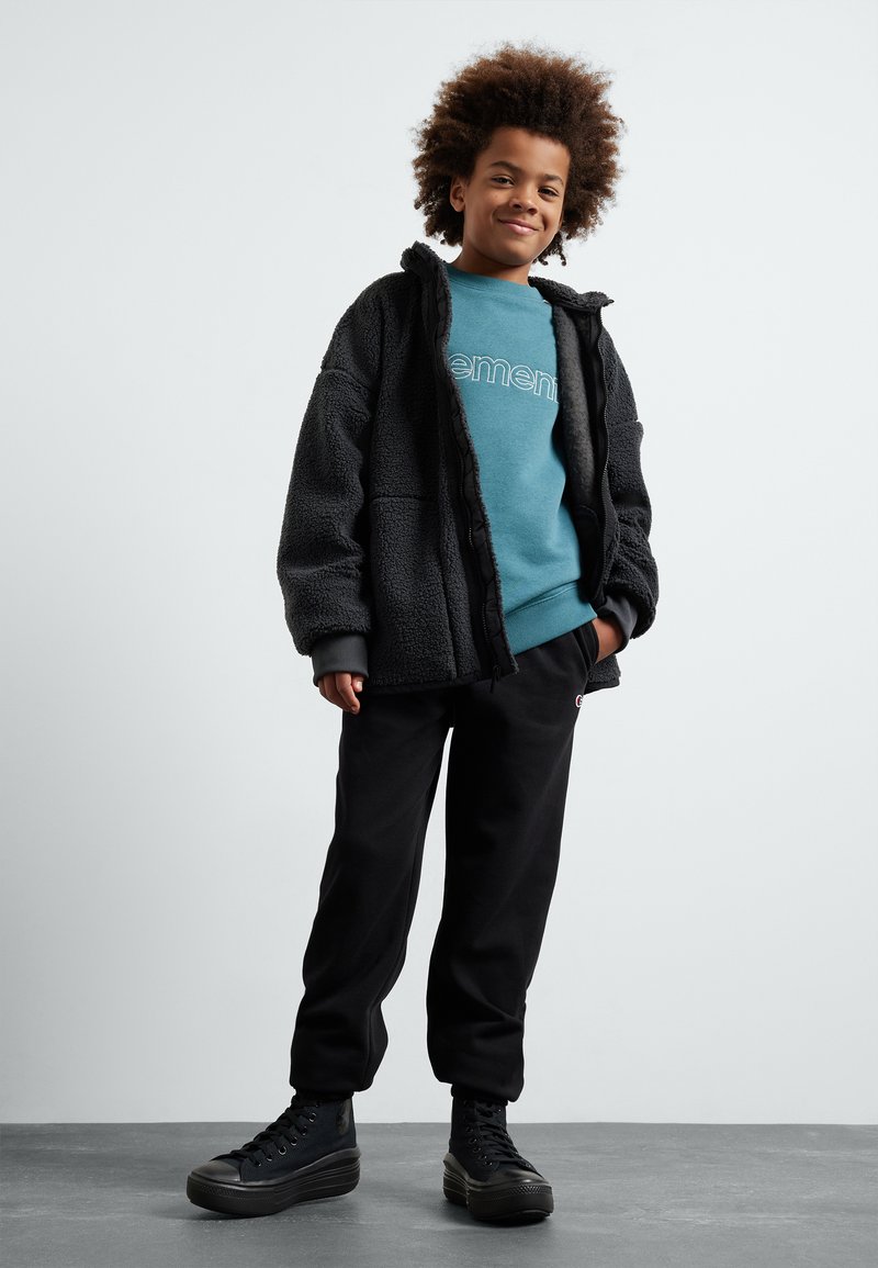 Veste en polaire noire avec un aspect texturé, sweat-shirt bleu avec logo, pantalon de survêtement noir et baskets montantes noires.