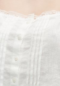 Κοντινό πλάνο λευκού μπλουζιού με κουμπιά, κατακόρυφα πιέτες και λεπτή δαντέλα κατά μήκος του λαιμού σε ανοιχτόχρωμο δέρμα.