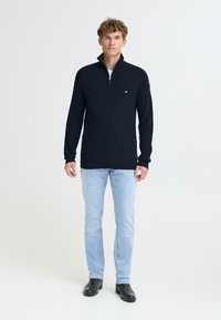 Pullover in maglina blu navy con zip a quarto, texture a coste e dettaglio del logo, abbinato a jeans blu chiaro e stivali neri alla caviglia.