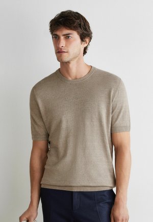 Ung mand med mørkt hår iført en beige kortærmet sweater og mørke bukser, stående mod en ensfarvet lys væg.