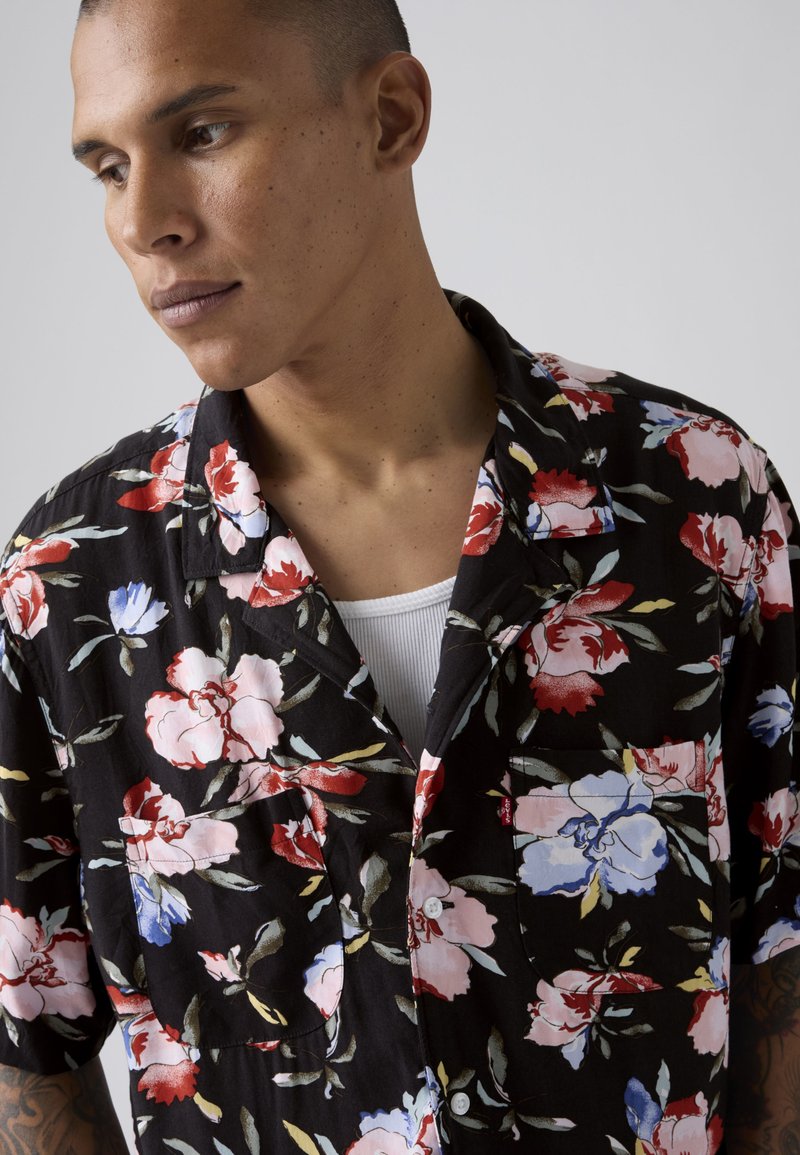 Ung mand med kort hår har en sort blomster skjorte over en hvid ribbet tanktop og kigger let ned mod venstre mod en grå baggrund.