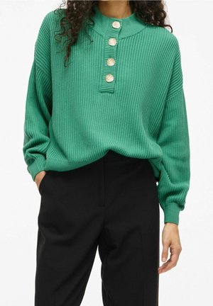 Femme portant un pull côtelé vert avec quatre grands boutons beige et un pantalon noir taille haute, mains détendues, une dans la poche.