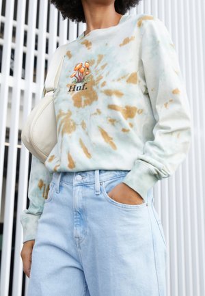 Persoon draagt lichtblauwe en beige tie-dye trui met paddenstoelenontwerp en de tekst "Huf", lichtblauwe spijkerbroek en witte schoudertas.