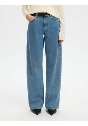 Persoon draagt lichtblauwe, hooggetailleerde brede jeans met een zwarte riem en puntige zwarte schoenen, handen gedeeltelijk in de zakken.