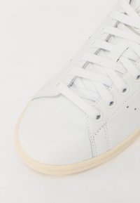 Zapatilla de cuero blanca con puntera redonda, cordones blancos y detalles de costura sutiles. Suela de goma con un acento en beige claro.