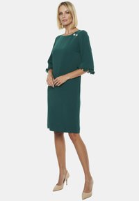 Sukienka w kolorze leśnej zieleni z rękawami o długości 3/4, z falbaniastymi brzegami, prostym krojem i dwoma dekoracyjnymi guzikami na lewym ramieniu.