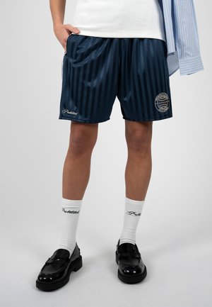 Person iført navy stribede shorts med logoer, hvide mærkede sokker, sorte loafers og en indstoppet hvid tanktop med en blå stribet skjorte draperet over den ene skulder.