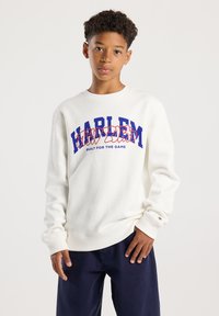 Niño con cabello rizado que lleva un sudadera blanca de Harlem y pantalones azul oscuro, de pie frente a un fondo claro y liso.