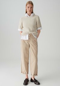 Maglione beige chiaro con maniche corte, sovrapposto a una camicia bianca con colletto. Pantaloni in corduroy beige con risvolti. Mocassini marroni scuri.