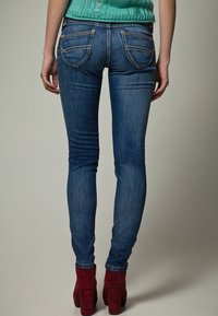 Hubené džíny v středně modrém praní s detaily na zadních kapsách, kombinované s červenými platformovými podpatky, z pohledu zezadu.