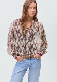 Blouse fluide multicolore avec un motif ikat, manches longues et décolleté en V, associée à un jeans bleu et une ceinture marron.
