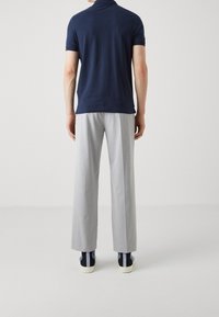 Mann in navyblauem Poloshirt, hellgrauen Hose und blauen Turnschuhen steht mit dem Rücken zu einer schlichten hellen Hintergrund.