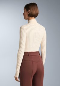 Krém színű bordázott kötött pulcsi magas gallérral, testhezálló fazonban és hosszú ujjakkal, rozsdabarna, magas derekú nadrággal párosítva.