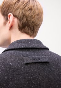 Dunkelgrauer Wollmantel mit breitem Kragen, strukturiertem Stoff mit kleinen Flecken und einem Markenpatch auf der Rückseite. Rückansicht des Nackens und der Haare des Models.