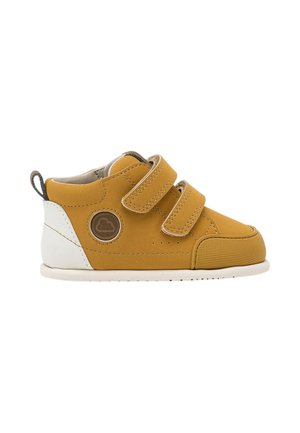 Zapato infantil amarillo mostaza con talón blanco, dos tiras de velcro, puntera de goma y logo circular en el lateral.