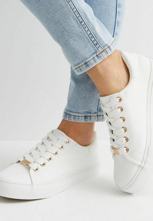 Sneakers bianchi in tela con una finitura testurizzata, dotati di occhielli dorati e una suola piatta in gomma, abbinati a jeans azzurri chiari con risvolto.