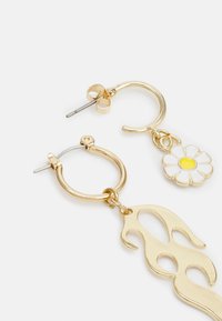 Brincos de argola dourados com um charme de margarida featuring pétalas brancas e um centro amarelo, e um pendente em forma de chama abstrata.