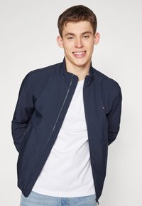 Giacca leggera blu navy con colletto rialzato e zip frontale, caratterizzata da cuciture sottili e un piccolo logo sul petto. Indossata sopra una t-shirt bianca.