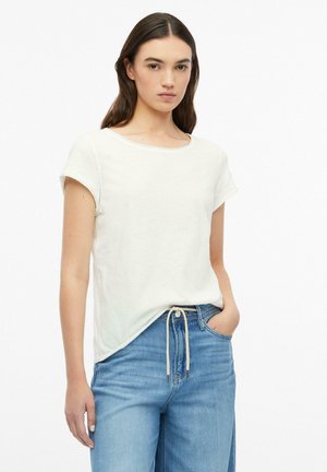 Vrouw met lang donker haar, die een wit T-shirt met korte mouwen en een blauwe spijkerbroek met een vastgeknoopt trekkoord draagt, staat met één hand in de zak.