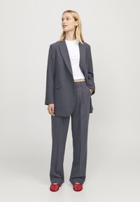 Grå skreddersydd blazer med lange ermer, kombinert med matchende bukser og en hvit crop top. Har røde perforerte slip-on sko.