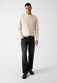 Sweat-shirt beige clair à col rond, jean en denim foncé, chaussures noires au design texturé. Le mannequin pose sur un fond neutre.