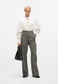 Weiße oversized Bluse mit "KARL"-Schriftzug, kombiniert mit hoch taillierten grauen Karohosen. Das Model hält eine schwarze strukturierte Handtasche.