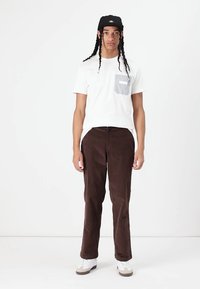 Tricou alb cu mânecă scurtă, având un buzunar în dungi, asortat cu pantaloni din catifea corduroy maro și adidași albi. Modelul are părul lung, împletit.