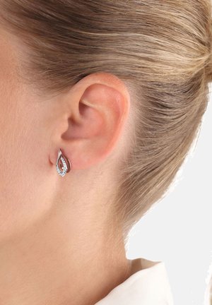 Primo piano di un orecchio femminile con capelli biondo chiaro tirati indietro, che indossa un piccolo orecchino a forma di foglia in argento con pietre trasparenti incastonate.