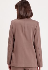 Blazer taillé marron avec un design minimaliste, une texture lisse et une coupe structurée, présentant des détails de coutures visibles et des manches longues.