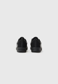 Ein Paar schwarze Sneakers, von hinten gezeigt, mit Zugschlaufen und flachen Gummisohlen, vor einem schlichten hellen Hintergrund.