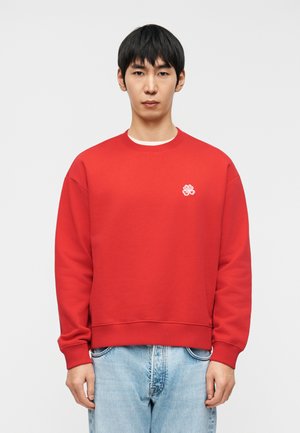 Jeune homme aux cheveux noirs portant un sweat-shirt rouge uni avec un petit emblème blanc et un jean bleu clair, debout devant un fond blanc.