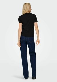 Kvinde med langt lyst hår står med ryggen til, iført en sort t-shirt med korte ærmer, mørkeblå jeans og sorte højhælede sko på en ensfarvet baggrund.