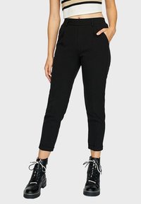 Pantalon noir ajusté avec une taille haute, des poches latérales et des ourlets roulés. Porté avec des bottines à lacets noires.
