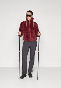 Fleecejacka i burgund med huva och snoddar, tillsammans med grå byxor och svarta vandringskängor, hållande vandringsstavar.