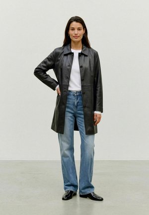 Une femme porte un manteau en cuir noir boutonné sur le devant, par-dessus une chemise blanche, un jean bleu et des chaussures noires, devant un mur de studio uni.