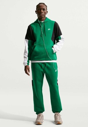 Person står iført en grøn, sort og hvid Nike hættetrøje og matchende bukser med beige sneakers og hvide høretelefoner.