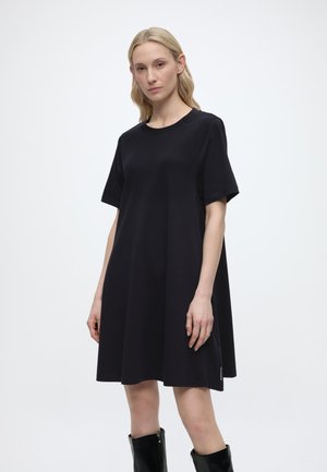 Blondiin naine, kes kannab lahtist musta lühikeste varrukatega kleiti ja põlveni ulatuvaid musti saapaid, valgel taustal.