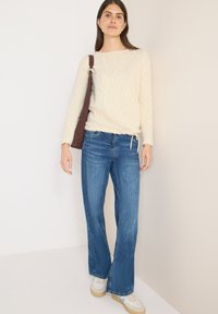 Suéter de punto texturizado color crema con patrón de ondas, combinado con vaqueros acampanados azules y zapatillas blancas. Bolso de hombro marrón con herrajes plateados.