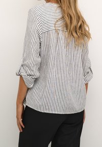 Blusa bianca e blu a righe con maniche arrotolate, dettaglio della piega sul retro e texture morbida, abbinata a pantaloni neri.