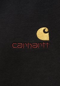 Μαύρο ύφασμα με κεντημένο λογότυπο Carhartt που περιλαμβάνει κίτρινο καμπύλο σχήμα και το όνομα της μάρκας με κόκκινη κλωστή.