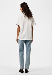 Person with curly hair facing away, wearing an oversized white t-shirt, light blue jeans, and black sneakers, standing on a white background.