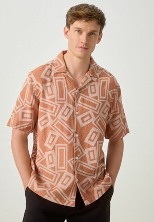 Junger Mann mit kurzem braunem Haar, der ein kurzärmliges, rostfarbenes Hemd mit hellen geometrischen Mustern trägt, die Hände in den Taschen.
