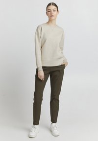 Beige strikkegenser med rund hals og ribbede mansjetter, kombinert med olivengrønne slim-fit bukser. Hvite sneakers fullfører antrekket.