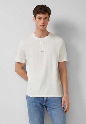 Junger Mann mit kurzen braunen Haaren, der ein weißes kurzärmeliges Henley-Shirt und blaue Jeans trägt, steht vor einer grauen Wand.