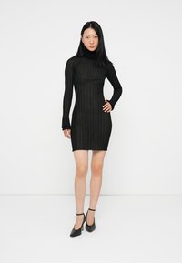 Μαύρο πλεκτό φόρεμα με μακριά μανίκια, ψηλό λαιμό, εφαρμοστή σιλουέτα και υφασμάτινο σχέδιο. Συνδυασμένο με μαύρες γόβες με λουρί στον αστράγαλο.