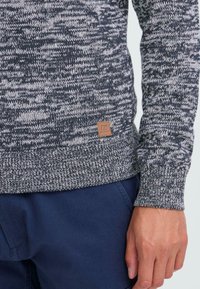Primer plano de una persona que lleva un suéter de punto jaspeado en gris y negro con un pequeño parche de logo marrón, combinado con pantalones azul marino.